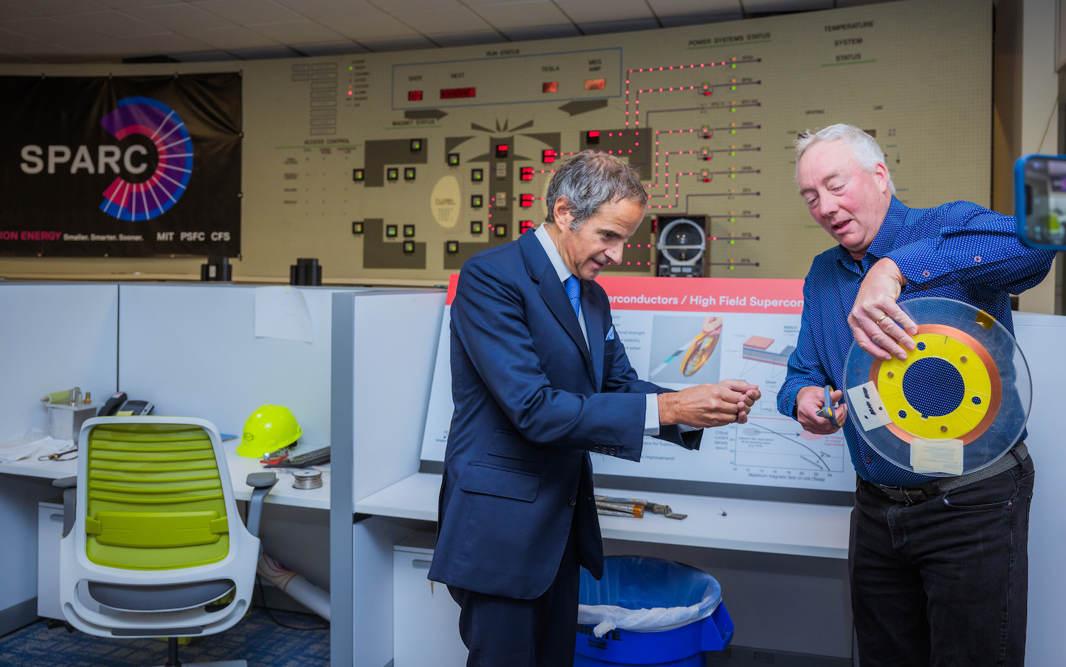 Dennis Whyte and Raphael Mariano Grossi stand in front of a retro-looking scientific control panel as Dennis shows Raphael a piece of high-temperature superconducting tape used in fusion magnets. 
