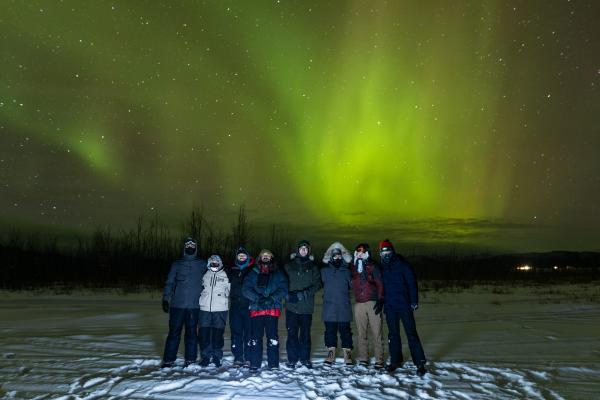 Group of 8 graduate students under a bright green aurora, filling the entire sky behind them