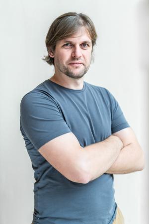 Headshot of Michael Komm; a white man with light brown hair and stubble. He has a blue t-shirt and his arms crossed, looking at the camera