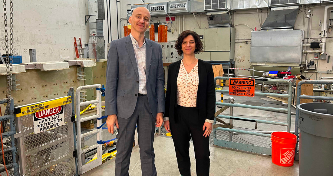 A man and a woman scientist stand next to one another and smile next to one another as they stand in front of a partially-deconstructed fusion device that is housed in a large concrete-lined room on MIT's campus.