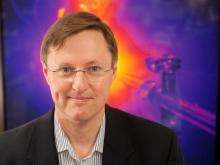 Johan Frenje against an image of a laser- the laser is visible with an orange center, fading to red and then purple. Johan is smiling in the left of the frame; a white man with short sandy hair, wire rimmed glasses, a dark jacket and a light gray button up shirt.