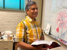 Abhay K Ram, a physics professor at MIT PSFC, smiles as he stands in front of a whiteboard holding a physics textbook. 